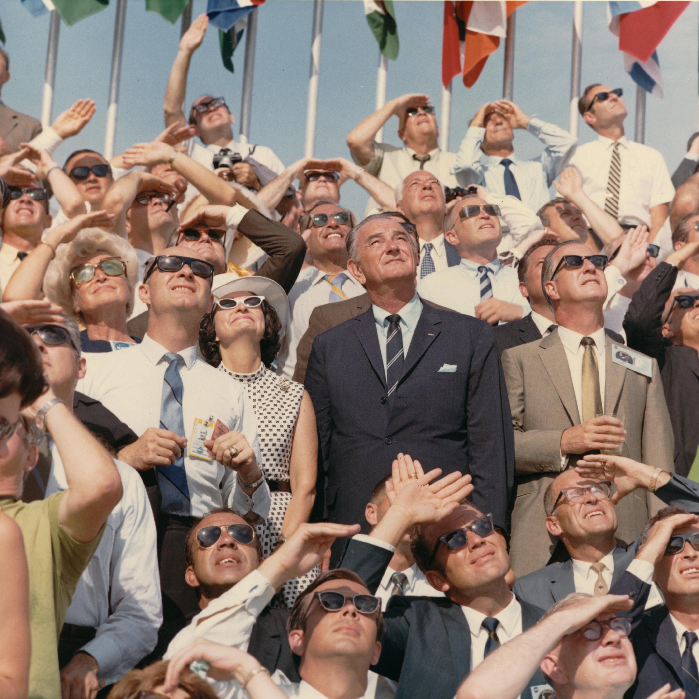 Lady Bird and Lyndon Johnson look to the sky amid a crowd of similar sky lookers, many with sunglasses.
